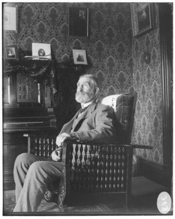 An elderly bearded man sitting in an arm chair. He is in a residence. surrounded by a piano. a trumpet. portrait photographs. and wall-