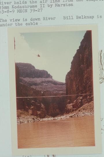 Lynn Roberts in his helicopter working as the elevator at the Marble Canyon Dam site. The cable across the river holds the air line from the compressors at the rim. The view is downriver. Bill Belknap is in his Sportyak under the cable